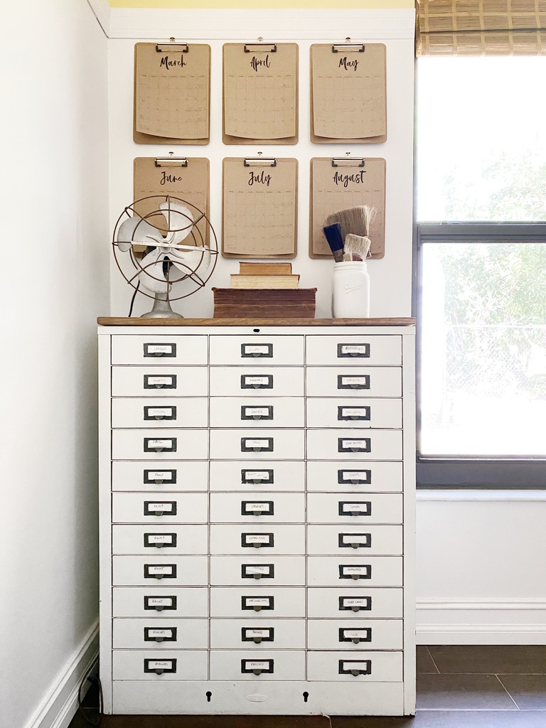 Vintage metal file drawers sit below a wall of clipboard calendars beside a light and airy window.