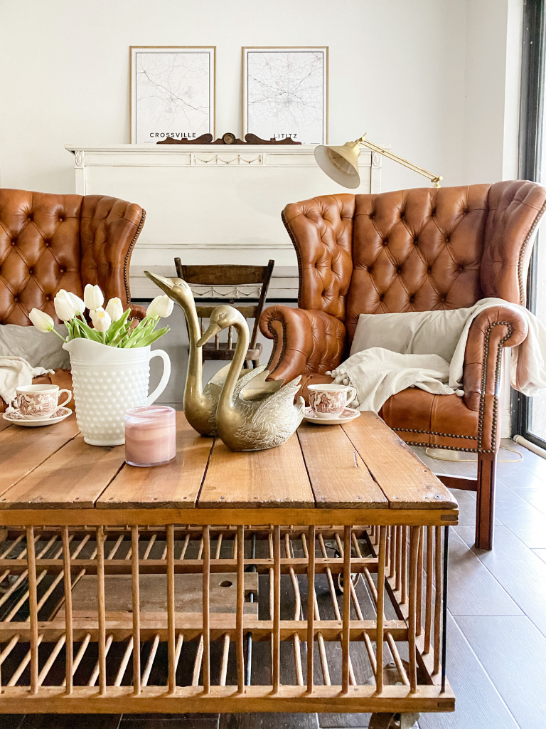 Two brown, tufted leather chairs sit behind a custom built chicken coop coffee table and in front of a painted white piano.